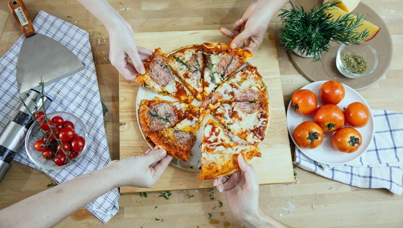Przyjaciele jedzą domową pizzę z salami, mozzarellą i sosem pomidorowym, na stole leżą pomidory i zioła.